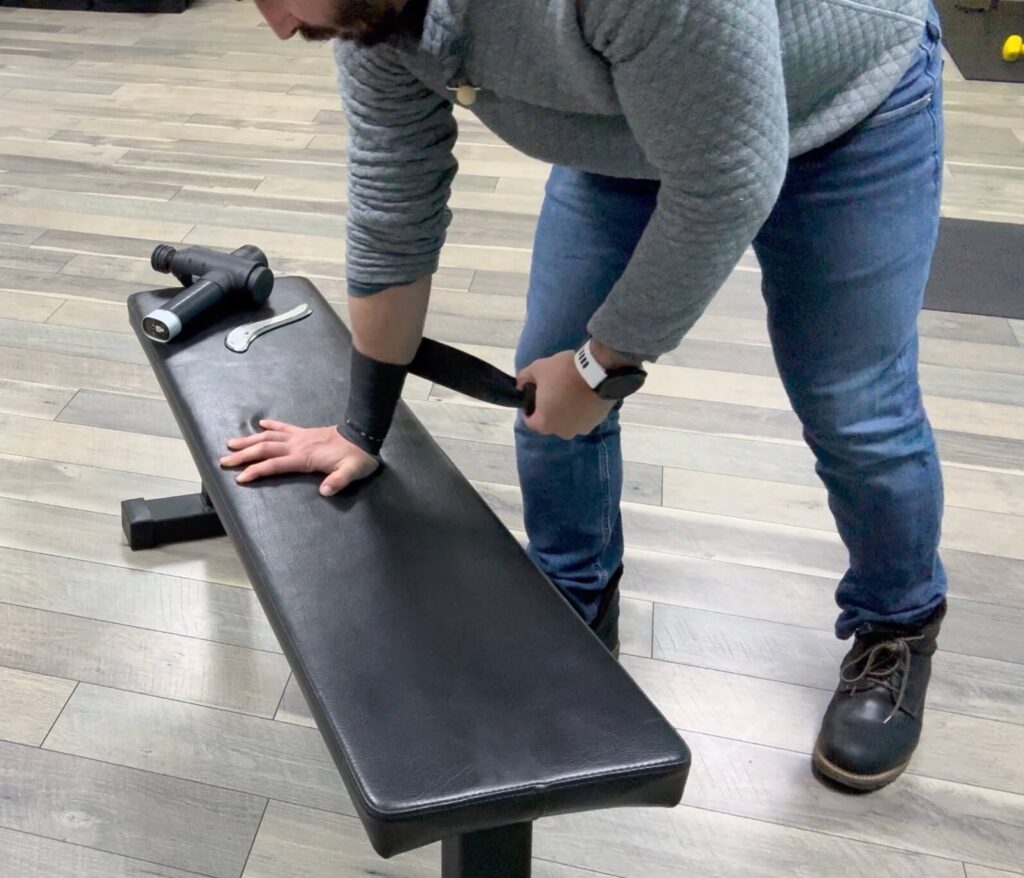 Dr. John with his right wrist wrapped with muscle floss. He is using a bench to apply pressure into the wrapped wrist to improve wrist pain by working on wrist extension.